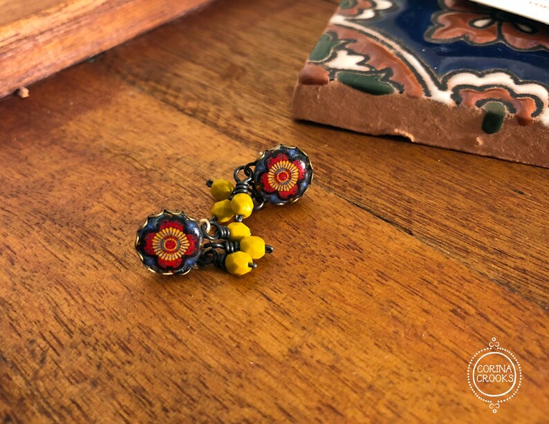 May include: Close-up of handmade earrings with a floral design. The earrings feature a red and yellow flower pattern set in a dark blue frame. Yellow beads dangle below the floral design. The earrings are on a wooden surface.