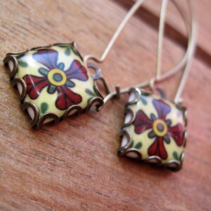 May include: A pair of square earrings with a floral design. The earrings feature a cream-colored background with a red and yellow flower design. The edges are framed with a bronze-colored metal. The earrings hang from thin, silver-colored wires.