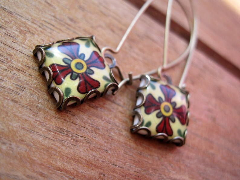 May include: A pair of square earrings with a floral design. The earrings feature a cream-colored background with a red and yellow flower design. The edges are framed with a bronze-colored metal. The earrings hang from thin, silver-colored wires.