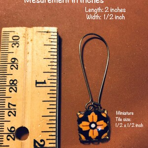 K&ouml;nnte beinhalten: Ein Paar goldfarbene Ohrringe mit einer schwarzen und orangefarbenen Fliese mit einem wei&szlig;en Blumenmuster. Die Fliese misst 1,27 cm x 1,27 cm. Die Ohrringe sind 5,08 cm lang.