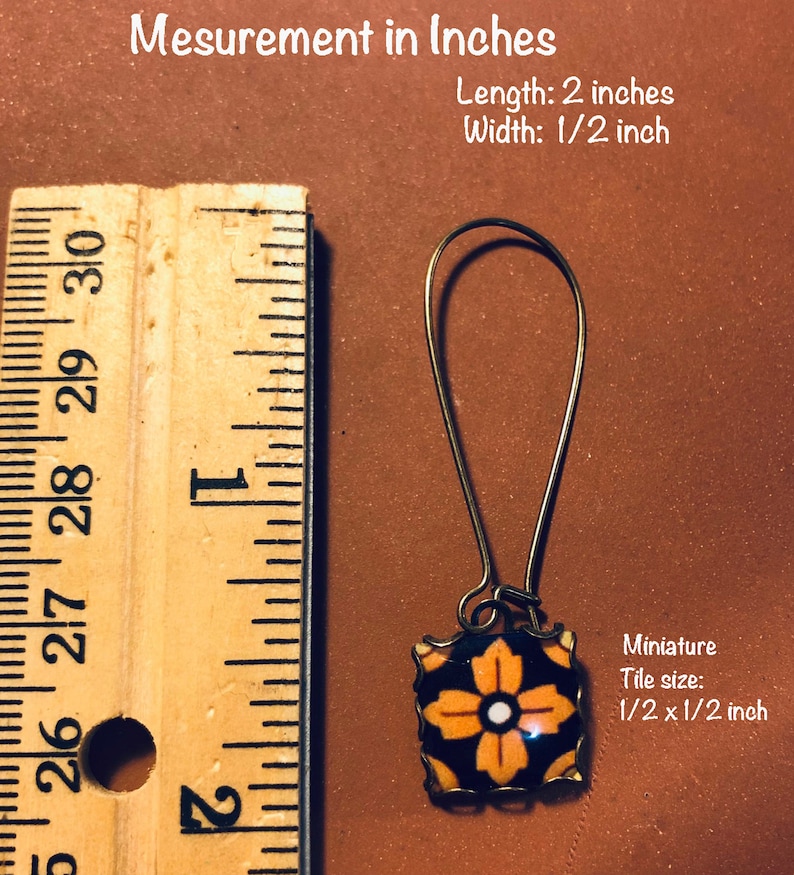 K&ouml;nnte beinhalten: Ein Paar goldfarbene Ohrringe mit einer schwarzen und orangefarbenen Fliese mit einem wei&szlig;en Blumenmuster. Die Fliese misst 1,27 cm x 1,27 cm. Die Ohrringe sind 5,08 cm lang.