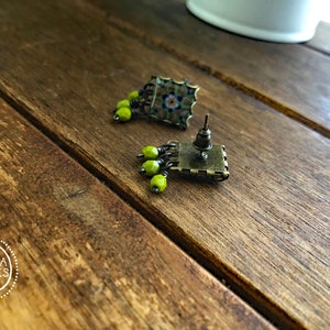 Pu&ograve; includere: Orecchini a bottone quadrati con un design a mosaico nei toni del verde, blu e marrone. Ogni orecchino presenta un gruppo di piccole perline rotonde verde lime. Finitura in ottone antico.