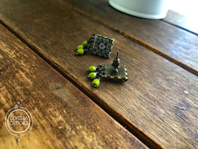 Pu&ograve; includere: Orecchini a bottone quadrati con un design a mosaico nei toni del verde, blu e marrone. Ogni orecchino presenta un gruppo di piccole perline rotonde verde lime. Finitura in ottone antico.