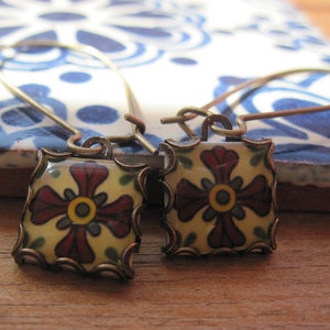 May include: A pair of square earrings with a floral design. The earrings feature a cream-colored background with a red and yellow floral pattern. The edges are framed with a bronze-colored metal. The earrings are on a blue and white patterned surface.