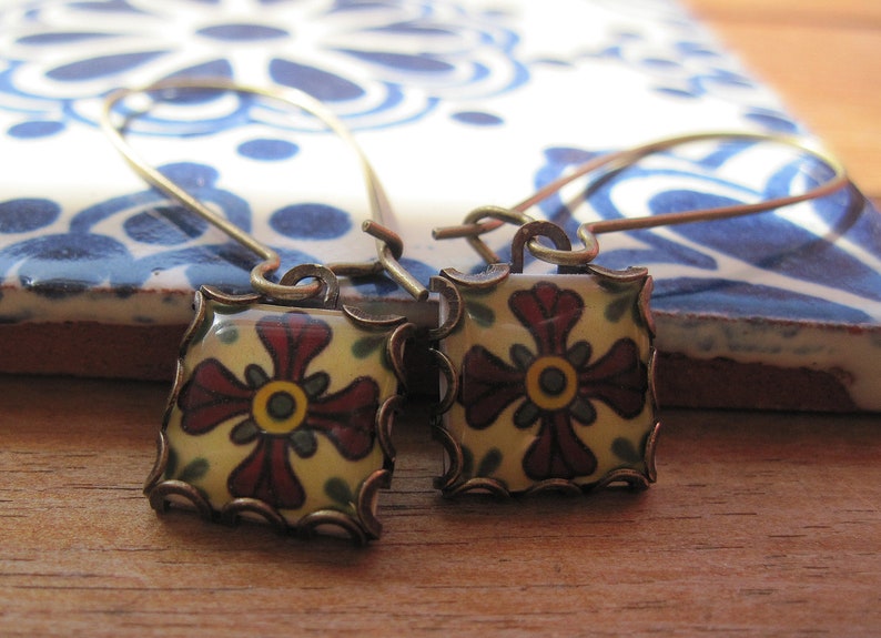 May include: A pair of square earrings with a floral design. The earrings feature a cream-colored background with a red and yellow floral pattern. The edges are framed with a bronze-colored metal. The earrings are on a blue and white patterned surface.