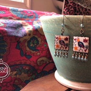 May include: Square earrings with a bird and floral design. The earrings have a white background with a colorful bird, orange flowers, and red flowers. The earrings have a bronze-colored frame and blue bead accents.