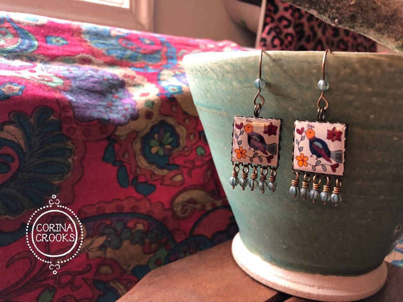 May include: Square earrings with a bird and floral design. The earrings have a white background with a colorful bird, orange flowers, and red flowers. The earrings have a bronze-colored frame and blue bead accents.