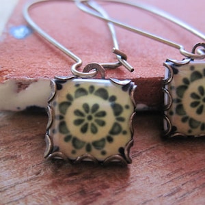 May include: A pair of square earrings with a floral design. The earrings feature a cream-colored background with a dark green floral pattern. The edges are framed with a bronze-colored metal. The earrings hang from a thin, silver-colored wire.