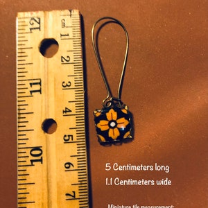 K&ouml;nnte beinhalten: Goldfarbene Ohrringe mit einem quadratischen Fliesenanh&auml;nger in Schwarz und Orange. Die Fliese hat ein wei&szlig;es Blumenmuster. Die Ohrringe sind 5 cm lang und 1,1 cm breit. Die Fliese misst 10 mm mal 10 mm.