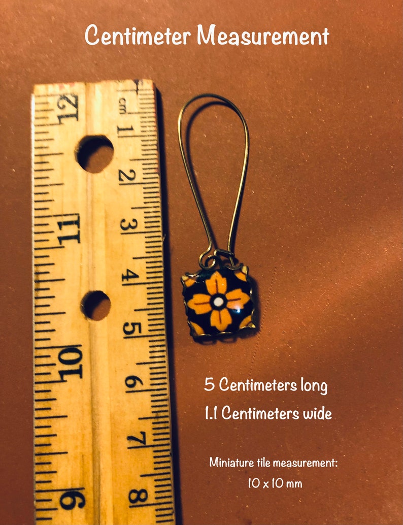 K&ouml;nnte beinhalten: Goldfarbene Ohrringe mit einem quadratischen Fliesenanh&auml;nger in Schwarz und Orange. Die Fliese hat ein wei&szlig;es Blumenmuster. Die Ohrringe sind 5 cm lang und 1,1 cm breit. Die Fliese misst 10 mm mal 10 mm.