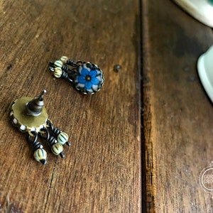 May include: A pair of antique-style earrings with a floral design. The earrings feature a blue flower with white accents and small, dangling beads. The metal has a dark, aged finish, and the earrings are set against a wooden surface.