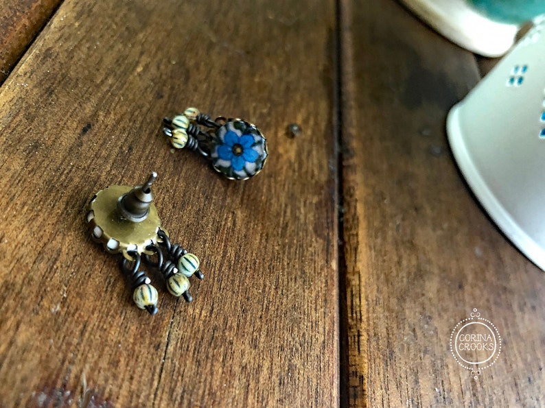 May include: A pair of antique-style earrings with a floral design. The earrings feature a blue flower with white accents and small, dangling beads. The metal has a dark, aged finish, and the earrings are set against a wooden surface.