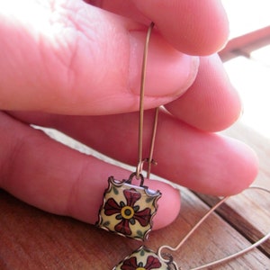 May include: A pair of square drop earrings with a floral design. The earrings feature a yellow background with a red flower and green leaves. The edges are outlined in a copper color. The earrings are held by a person.