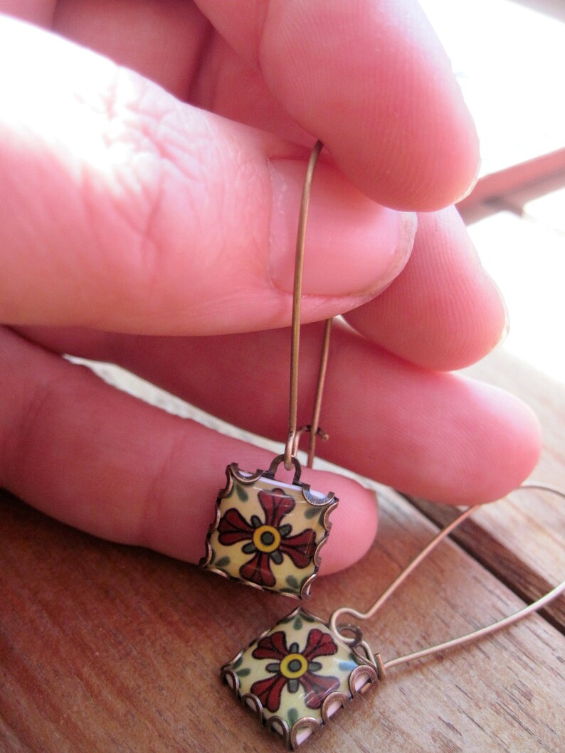 May include: A pair of square drop earrings with a floral design. The earrings feature a yellow background with a red flower and green leaves. The edges are outlined in a copper color. The earrings are held by a person.