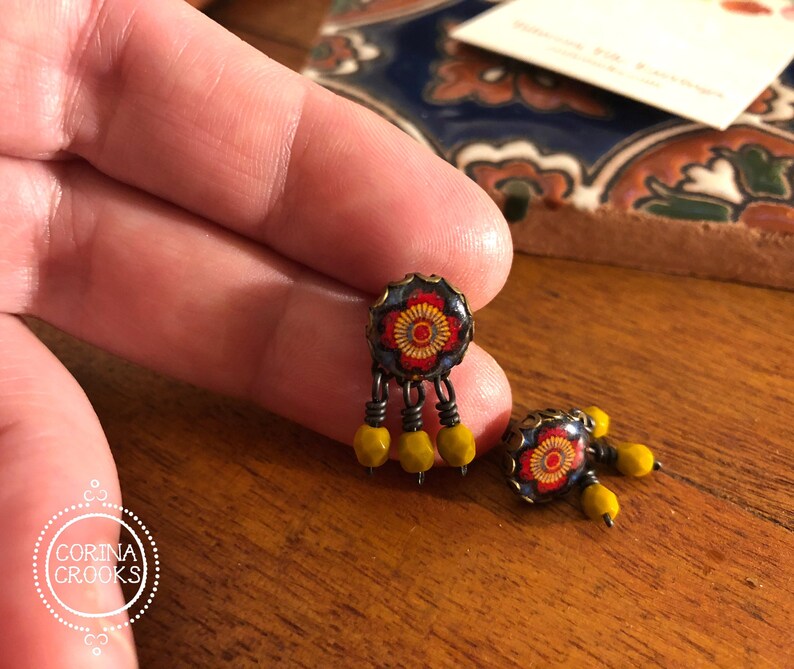 May include: A pair of handcrafted earrings featuring a floral design with red, yellow, and blue accents. The earrings have a dark metal frame and dangle with yellow beads. The earrings are displayed on a wooden surface.