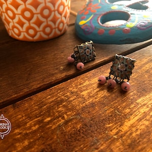 May include: A pair of blue and white square earrings with pink beads hanging from them. The earrings are on a wooden surface.