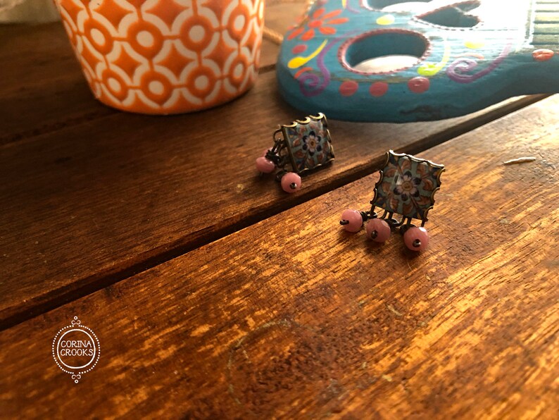 May include: A pair of blue and white square earrings with pink beads hanging from them. The earrings are on a wooden surface.