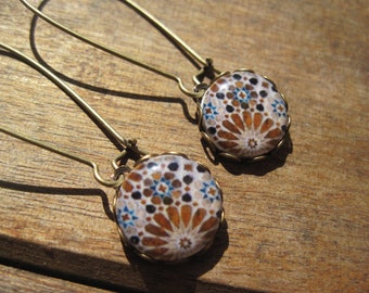 Può includere: Un paio di orecchini pendenti con un design a mosaico. Gli orecchini presentano pendenti rotondi e decorati nei toni del marrone, blu e bianco, incastonati in una montatura in metallo color oro. Gli orecchini hanno lunghi e sottili fili per le orecchie.