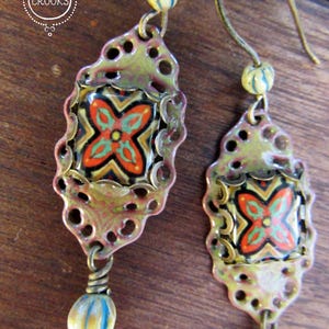 May include: A pair of handcrafted earrings with a floral design. The earrings feature a decorative metal frame in shades of green and pink, with a colorful floral pattern in the center. Each earring has a dangling bead with blue and yellow stripes.