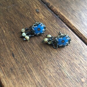 May include: A pair of handcrafted earrings featuring a blue floral design with a cream-colored background. The earrings have a dark metal wire frame and are accented with small, striped beads. The earrings are displayed on a wooden surface.