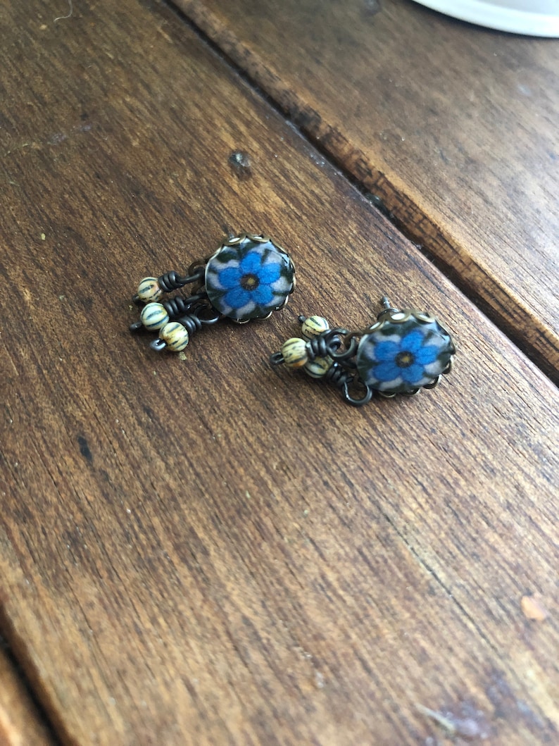 May include: A pair of handcrafted earrings featuring a blue floral design with a cream-colored background. The earrings have a dark metal wire frame and are accented with small, striped beads. The earrings are displayed on a wooden surface.