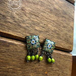 Pu&ograve; includere: Orecchini quadrati con motivo floreale verde, blu e bianco. Hanno una cornice color ottone e perline pendenti verde lime. Presentati su una superficie di legno.