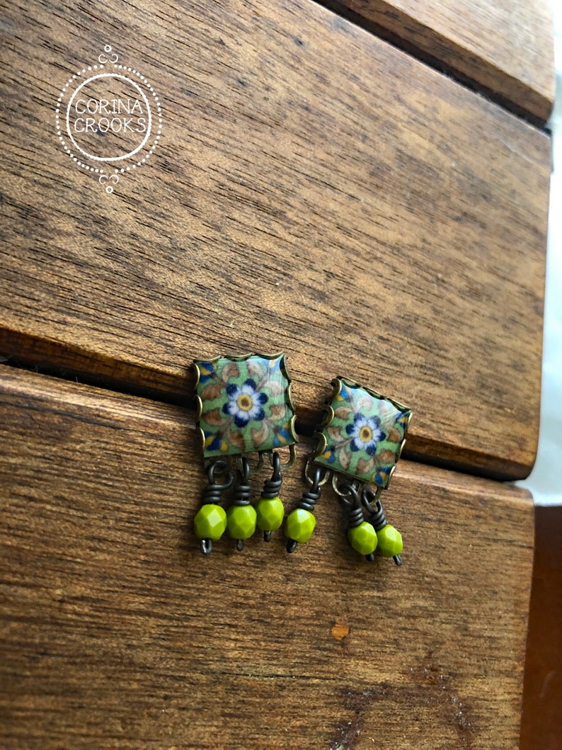 Pu&ograve; includere: Orecchini quadrati con motivo floreale verde, blu e bianco. Hanno una cornice color ottone e perline pendenti verde lime. Presentati su una superficie di legno.
