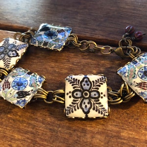 May include: A bracelet with square ceramic tiles, each displaying a detailed blue and white floral design. The tiles are linked by a gold-coloured chain. The bracelet fastens with a lobster clasp and has small red beads.