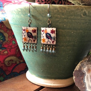 May include: A pair of square earrings with a bird design. The earrings feature a white background with a colorful bird, flowers, and a beaded fringe. The earrings are displayed on a green ceramic pot.