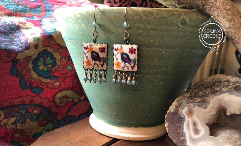 May include: A pair of square earrings with a bird design. The earrings feature a white background with a colorful bird, flowers, and a beaded fringe. The earrings are displayed on a green ceramic pot.