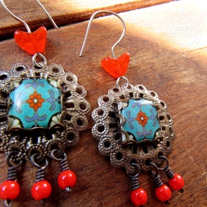 May include: A pair of dangle earrings with intricate details. Each earring features a square, turquoise tile with a floral design, set within a filigree frame. Red heart-shaped beads and dangling red beads complete the design. The earrings have silver-colored ear wires.