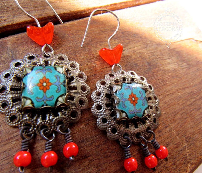 May include: A pair of dangle earrings with intricate details. Each earring features a square, turquoise tile with a floral design, set within a filigree frame. Red heart-shaped beads and dangling red beads complete the design. The earrings have silver-colored ear wires.