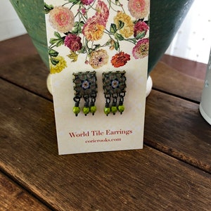 Pu&ograve; includere: Un paio di orecchini World Tile su una carta a motivi floreali. Gli orecchini hanno un design a piastrella quadrata con un motivo floreale e tre perline verdi appese a ciascun orecchino. La carta recita "World Tile Earrings" e "coricrooks.com".