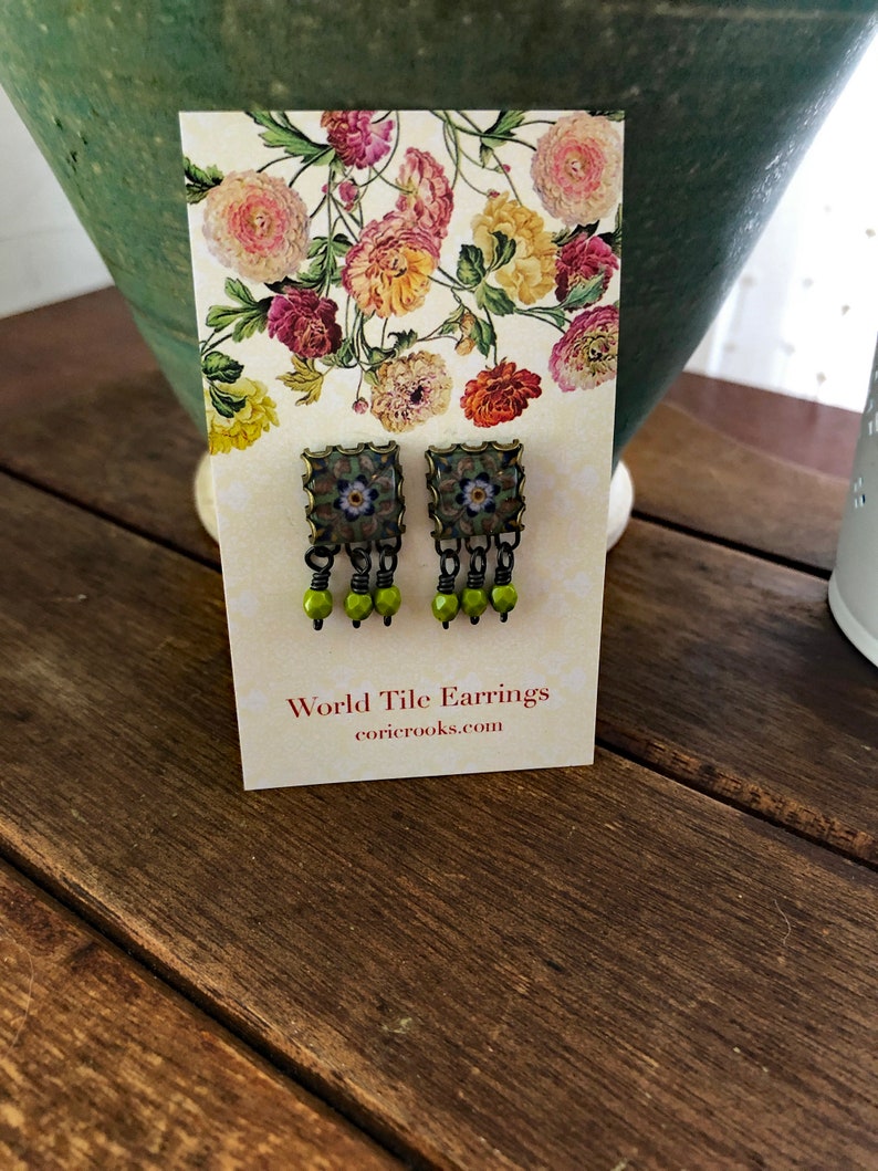 Pu&ograve; includere: Un paio di orecchini World Tile su una carta a motivi floreali. Gli orecchini hanno un design a piastrella quadrata con un motivo floreale e tre perline verdi appese a ciascun orecchino. La carta recita "World Tile Earrings" e "coricrooks.com".