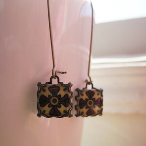 May include: A pair of square drop earrings with a floral design. The earrings feature a bronze-colored metal frame with a cream-colored center and a dark red and yellow floral pattern. The earrings hang from bronze-colored ear wires.