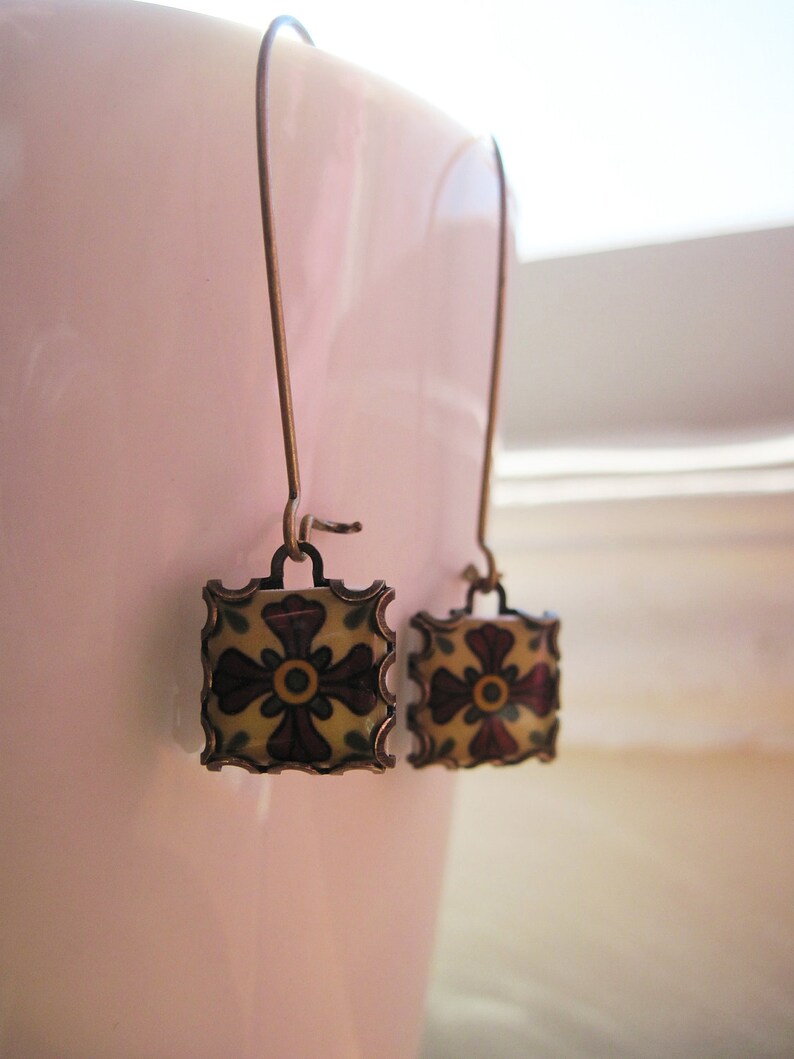 May include: A pair of square drop earrings with a floral design. The earrings feature a bronze-colored metal frame with a cream-colored center and a dark red and yellow floral pattern. The earrings hang from bronze-colored ear wires.