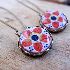 May include: A pair of earrings featuring a floral design. Each earring has a round, white base with a red, yellow, and blue floral pattern. The design is set in a gold-colored, scalloped frame with a long, curved ear wire.