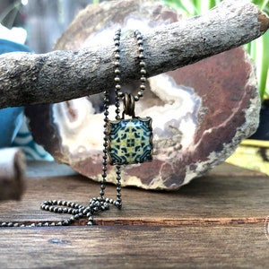 May include: A square pendant necklace with a blue and white patterned tile design. The pendant is attached to a silver chain.