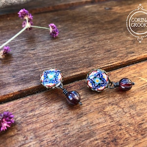 May include: A pair of handcrafted earrings featuring a square, colorful mosaic design with blue, red, and white accents. Each earring has a dangling, dark red bead. The earrings are set against a rustic wooden background, with the text "CORINA CROOKS" in a circle.
