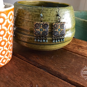 May include: A pair of square earrings with a floral design in shades of blue, gold, and teal. The earrings have a dark metal hook and dangle with small blue beads. The earrings are displayed near a green ceramic bowl and an orange and white patterned mug.