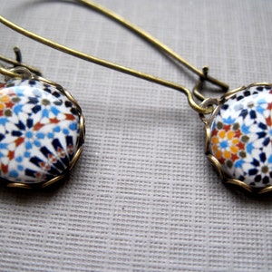 May include: A pair of dangle earrings with a mosaic design. The earrings feature round, white ceramic pendants with a colorful pattern of blue, orange, and brown shapes. The pendants are set in a gold-colored metal frame and hang from long, gold-colored ear wires.