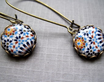 Puede incluir: Un par de pendientes colgantes con diseño de mosaico. Los pendientes presentan colgantes redondos de cerámica blanca con un patrón colorido de formas azules, naranjas y marrones. Los colgantes están engastados en un marco de metal dorado y cuelgan de largos ganchos dorados.