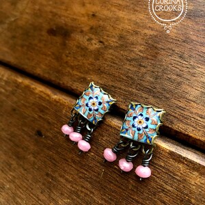 May include: A pair of blue and gold square earrings with pink beads hanging from the bottom. The earrings have a floral design.