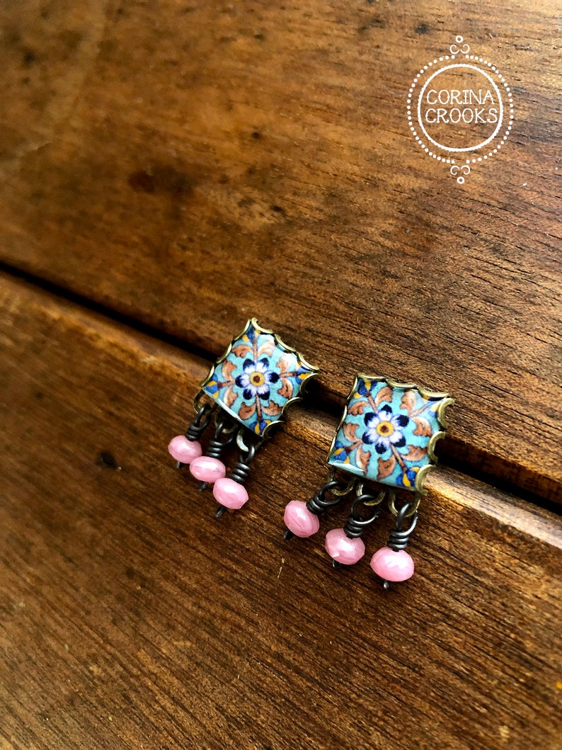 May include: A pair of blue and gold square earrings with pink beads hanging from the bottom. The earrings have a floral design.