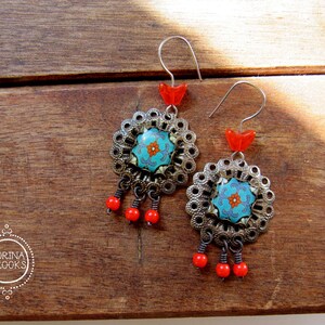 May include: A pair of ornate earrings with a floral design. The earrings feature a turquoise center with an orange flower, surrounded by a silver filigree frame. Red beads dangle below, and red accents are at the top. The earrings are on a wooden surface.
