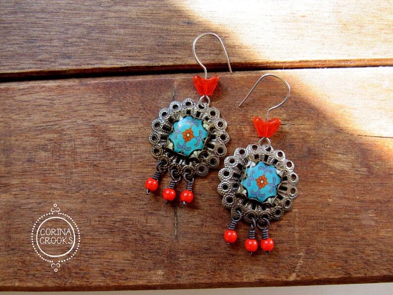May include: A pair of ornate earrings with a floral design. The earrings feature a turquoise center with an orange flower, surrounded by a silver filigree frame. Red beads dangle below, and red accents are at the top. The earrings are on a wooden surface.