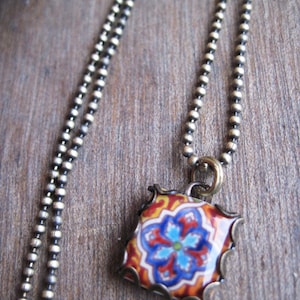 Puede incluir: Un collar de cadena plateada con un colgante cuadrado que presenta un diseño floral colorido. El colgante tiene un patrón rojo, naranja, azul y verde.