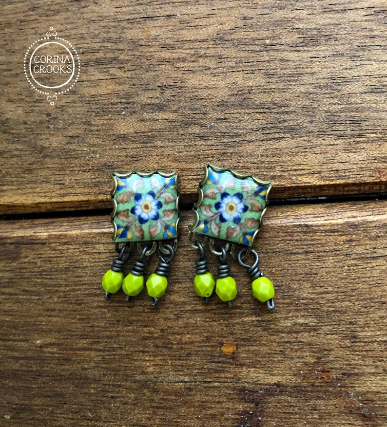 Pu&ograve; includere: Un paio di orecchini quadrati con un motivo floreale. Gli orecchini presentano uno sfondo verde chiaro con fiori blu e bianchi. Ogni orecchino ha tre perline pendenti verde neon. Gli orecchini sono appoggiati su una superficie di legno.
