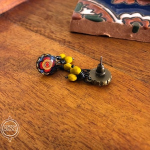 May include: A detailed earring with a floral design. The earring features a round, dark blue and red floral stud with yellow bead accents. The earring post is a bronze color. The earring is on a wooden surface.