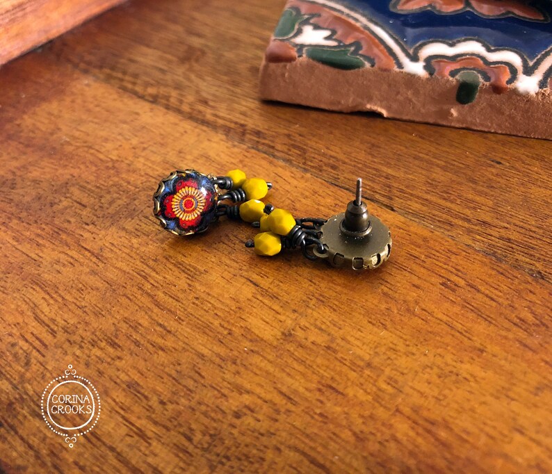 May include: A detailed earring with a floral design. The earring features a round, dark blue and red floral stud with yellow bead accents. The earring post is a bronze color. The earring is on a wooden surface.
